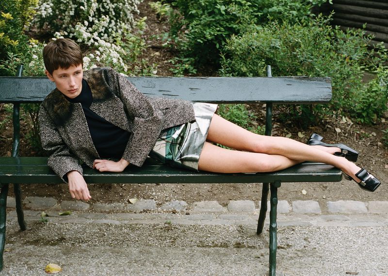 Woman in tweed jacket and metallic skirt reclines on a green park bench surrounded by lush greenery in an outdoor fashion shot.