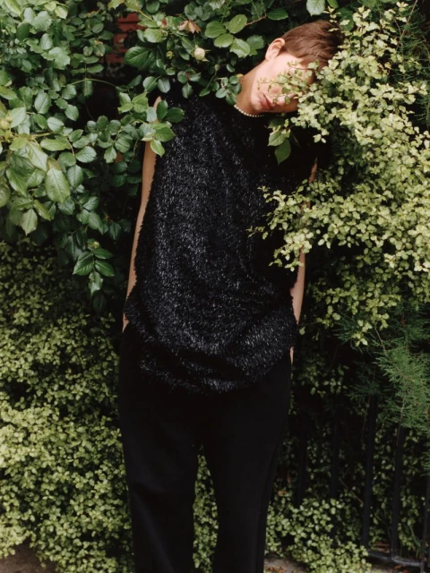 Adult wearing a black textured sleeveless top and black pants poses behind a green leafy bush in an outdoor setting.