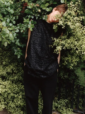 Adult wearing a black textured sleeveless top and black pants poses behind a green leafy bush in an outdoor setting.