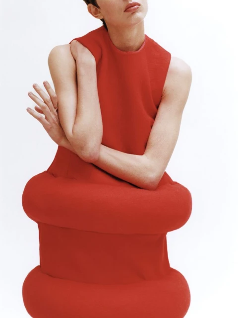 Woman in red dress with bulbous design poses against a white background, creating a striking fashion image.