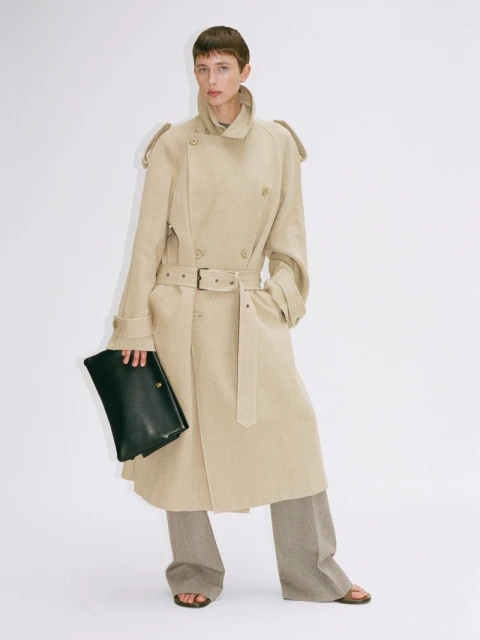 A young woman poses in a tan trench coat, light brown slacks, and sandals while holding a black leather clutch in a studio setting.