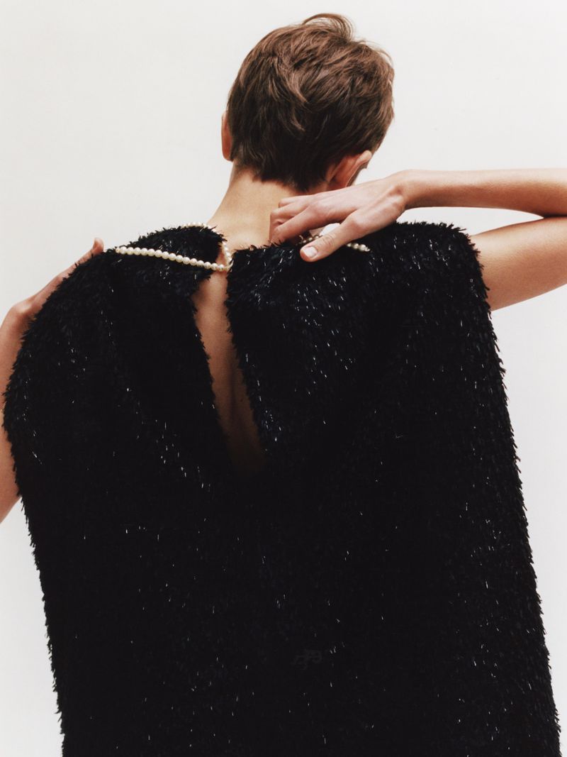 Back view of a woman in a black dress, adjusting a pearl necklace against a white background in a fashion setting.