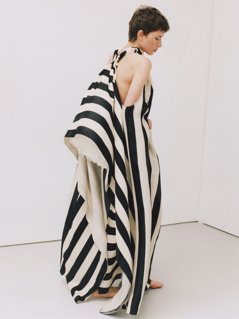 Adult woman in a black and white vertically striped sleeveless dress standing in a photo studio with bare feet and looking off camera.