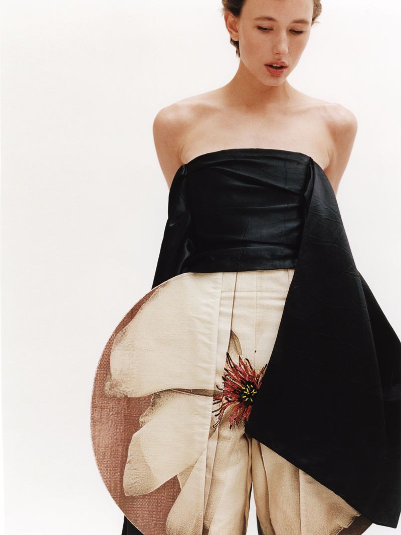 A fair-skinned woman poses in a strapless black and beige floral dress against a white backdrop, highlighting the dress's unique design and detail.