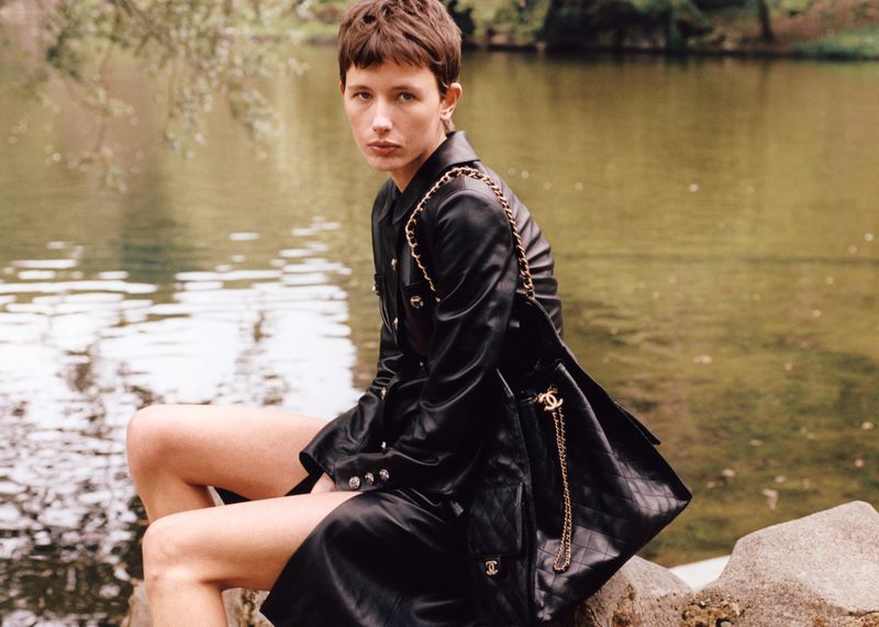 Female model in black leather trench coat and black shorts sitting on rocks next to a lake in a natural outdoor setting.