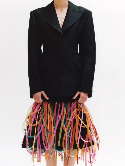 A fashion shot features an adult woman wearing a black blazer and a mini skirt adorned with colourful plastic tube accents, set against a white backdrop.