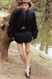 Model Nora Svenson wearing a black sculptural puffer top and mini skirt leaning against a tree trunk in a park.