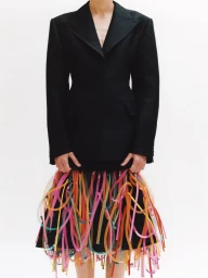 Model Nora Svenson wearing a black structured blazer and a vibrant multicolored tube fringe skirt against a plain white studio backdrop.