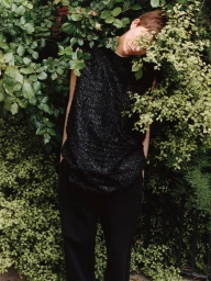 Model Nora Svenson leaning amidst dense green foliage wearing a textured black sleeveless top and dark trousers.