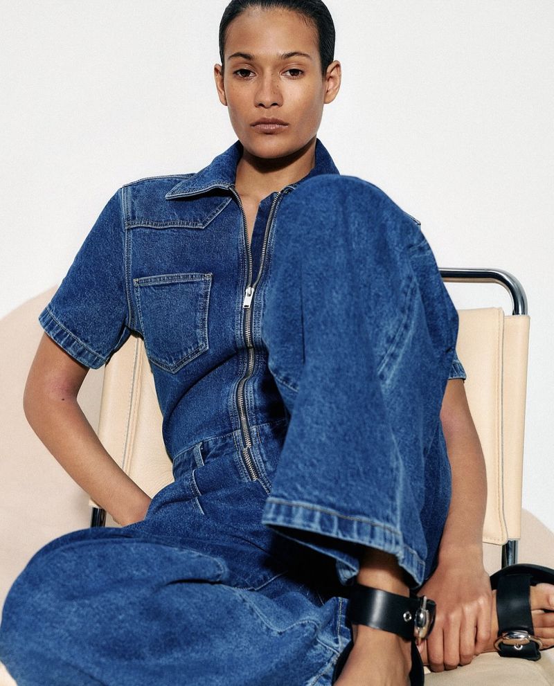 Fashion shot of a woman in a denim jumpsuit sitting on a chair against a white backdrop, showcasing a modern and casual style.