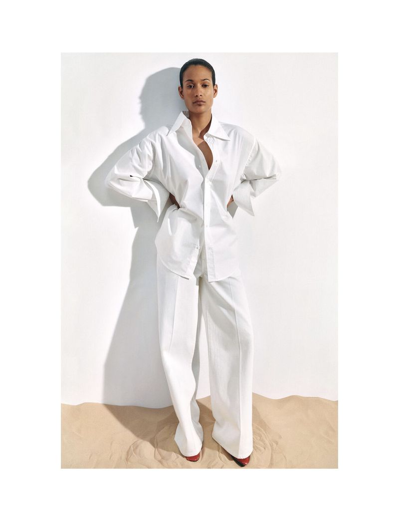 Fashion shot of a biracial woman in a white oversized shirt and pants, standing in front of a white wall and tan floor.