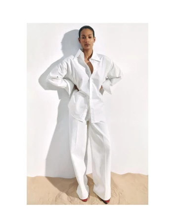 Fashion shot of a biracial woman in a white oversized shirt and pants, standing in front of a white wall and tan floor.