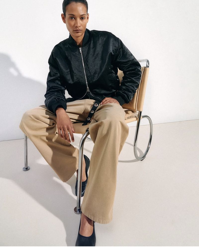 Fashion shot of a Black female model wearing a dark bomber jacket and beige trousers, seated on a beige chair against a white backdrop.