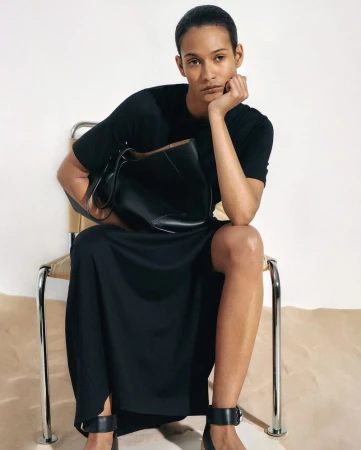 Adult female fashion model in black dress sits on a chair indoors, holding a large black purse, lit by soft studio lighting.