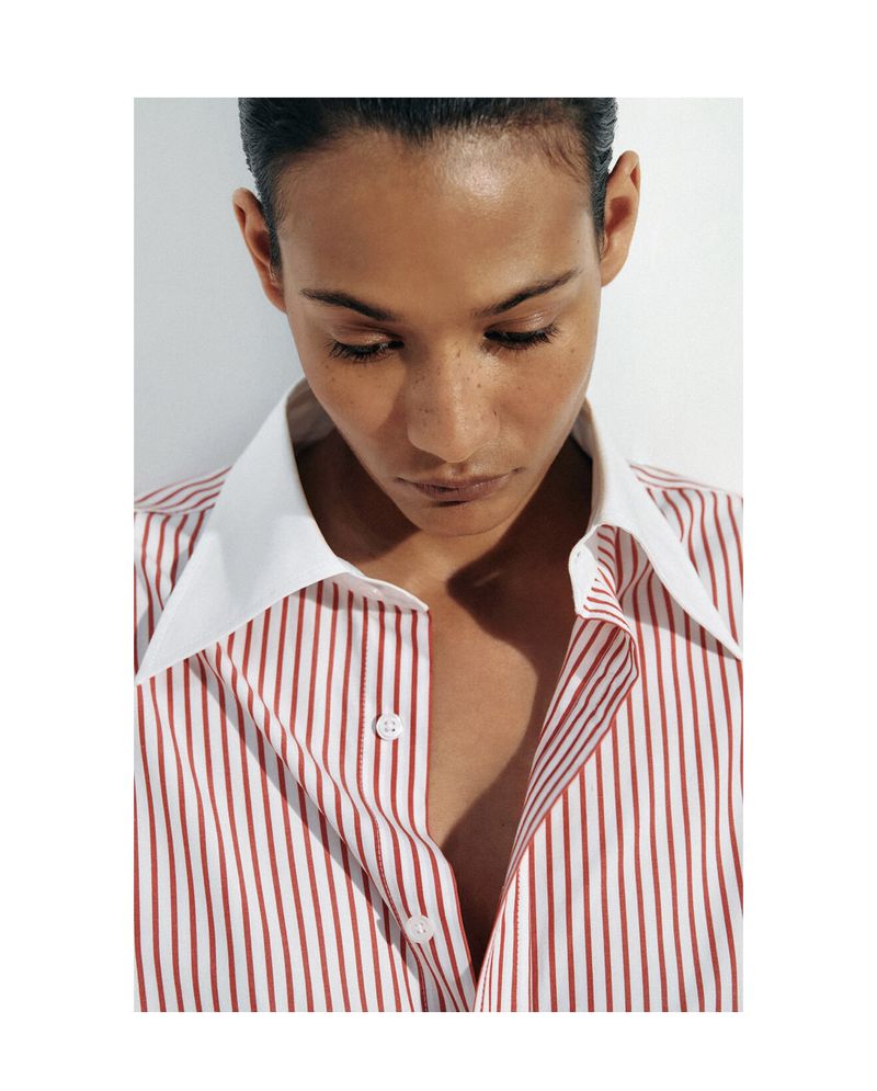Adult woman with tanned skin and short dark hair poses indoors, wearing a striped collared shirt against a plain white backdrop.