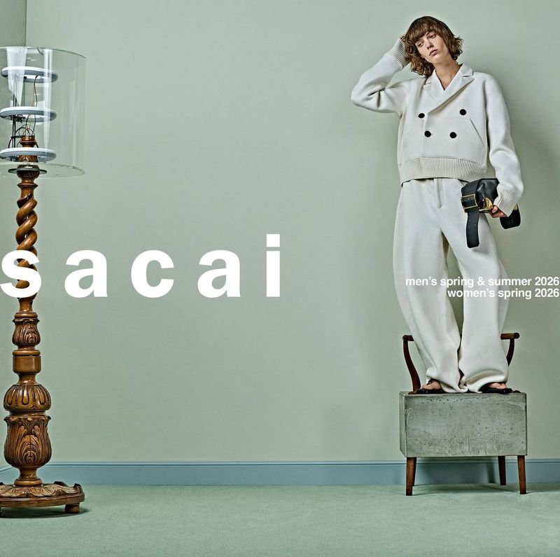 Editorial fashion shot of a person in a white suit standing on a concrete block, next to a wood lamp and Sacai logo.