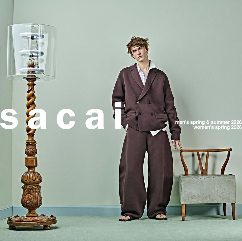 Fashion editorial featuring a stylish adult posing in a brown double-breasted suit next to a vintage lamp and chair in a studio.