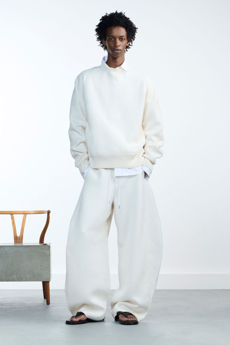 Full-length shot of adult male fashion model posing in oversized, off-white sweatshirt and pants, accessorized with sandals, against a white backdrop.