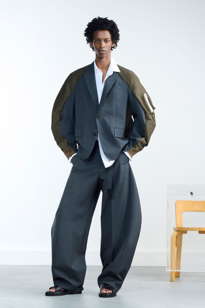 Full-length studio shot of an adult male model in a grey suit, open white dress shirt, olive jacket, black sandals, and natural hair.