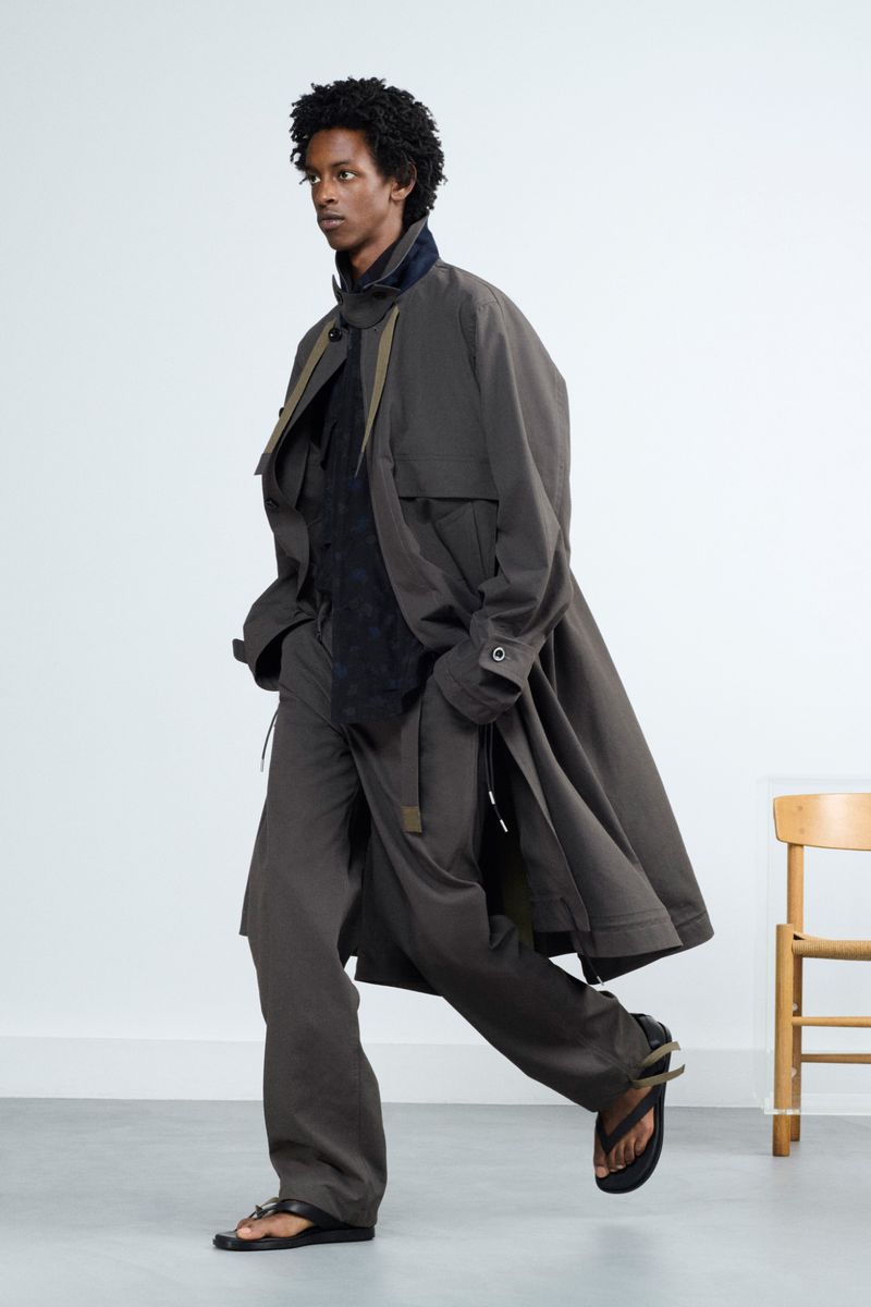 Fashionable man strides confidently in an olive-green trench coat and wide-leg pants, complemented by black sandals against a clean white background.