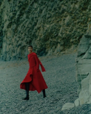 Model Nora Attal in a vibrant red draped dress walking across a dark pebble beach beneath a steep rocky cliff face.