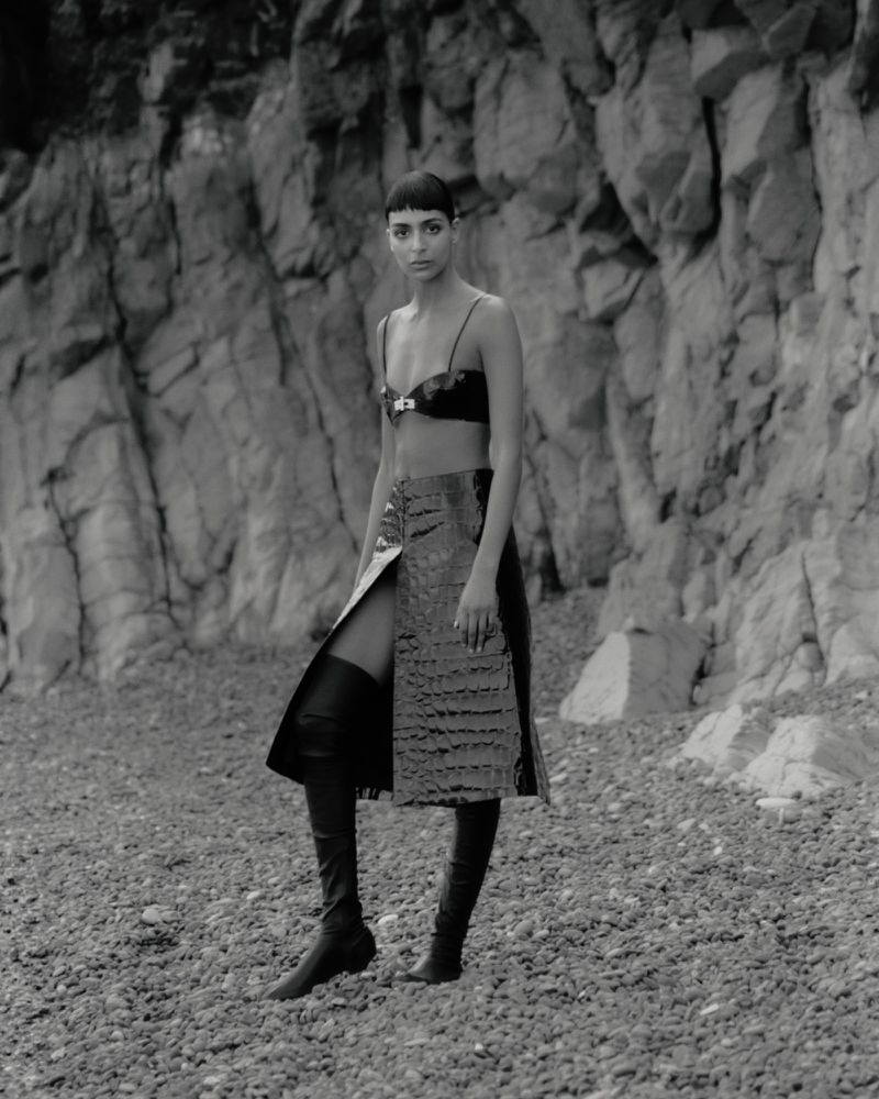A fashion model in a bralette, skirt, and knee-high boots poses on a rocky terrain in front of large rock formations in a studio shot.