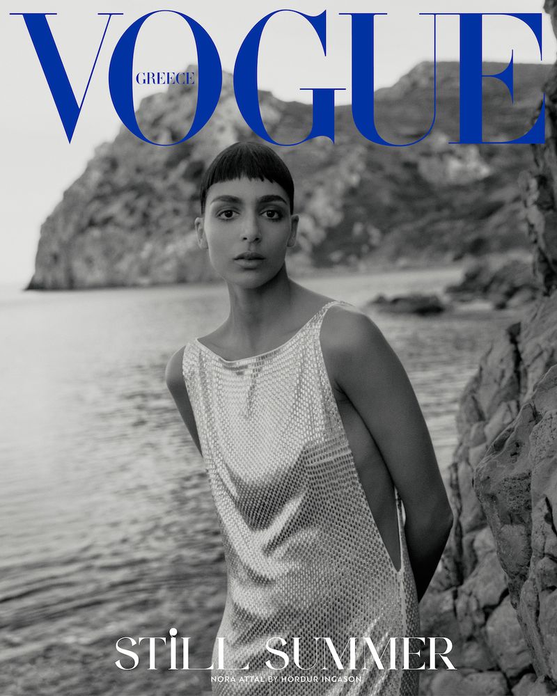 Vogue Greece magazine cover featuring a model in a shimmering dress on a rocky coast, with the ocean and sky in the background.