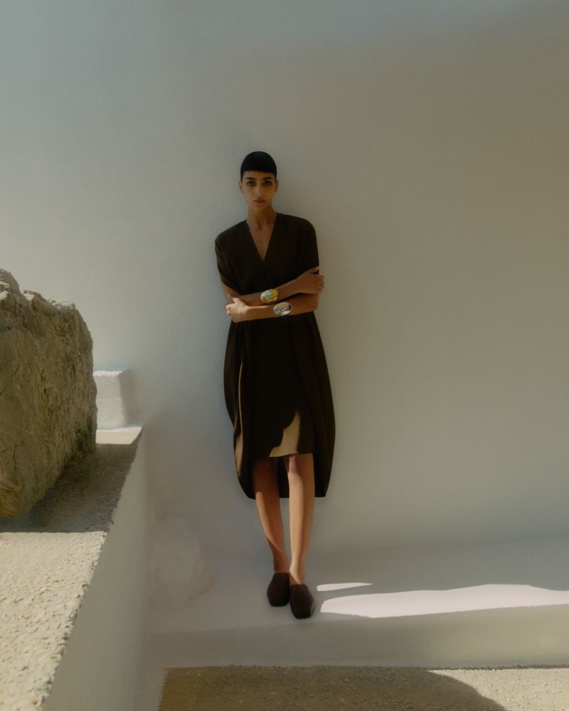 A brunette woman in a V-neck dress and flats stands against a white wall in a medium shot taken on a sunny day.