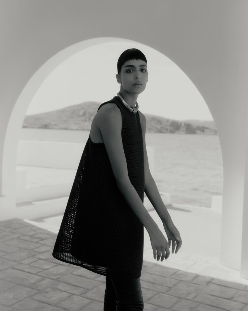 Model Nora Attal in a black sleeveless trapeze dress standing under an arched terrace overlooking the sea in black and white.