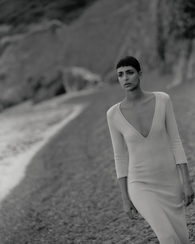 Model Nora Attal walking along a beach in a long deep V-neck knit dress in a black and white fashion editorial.