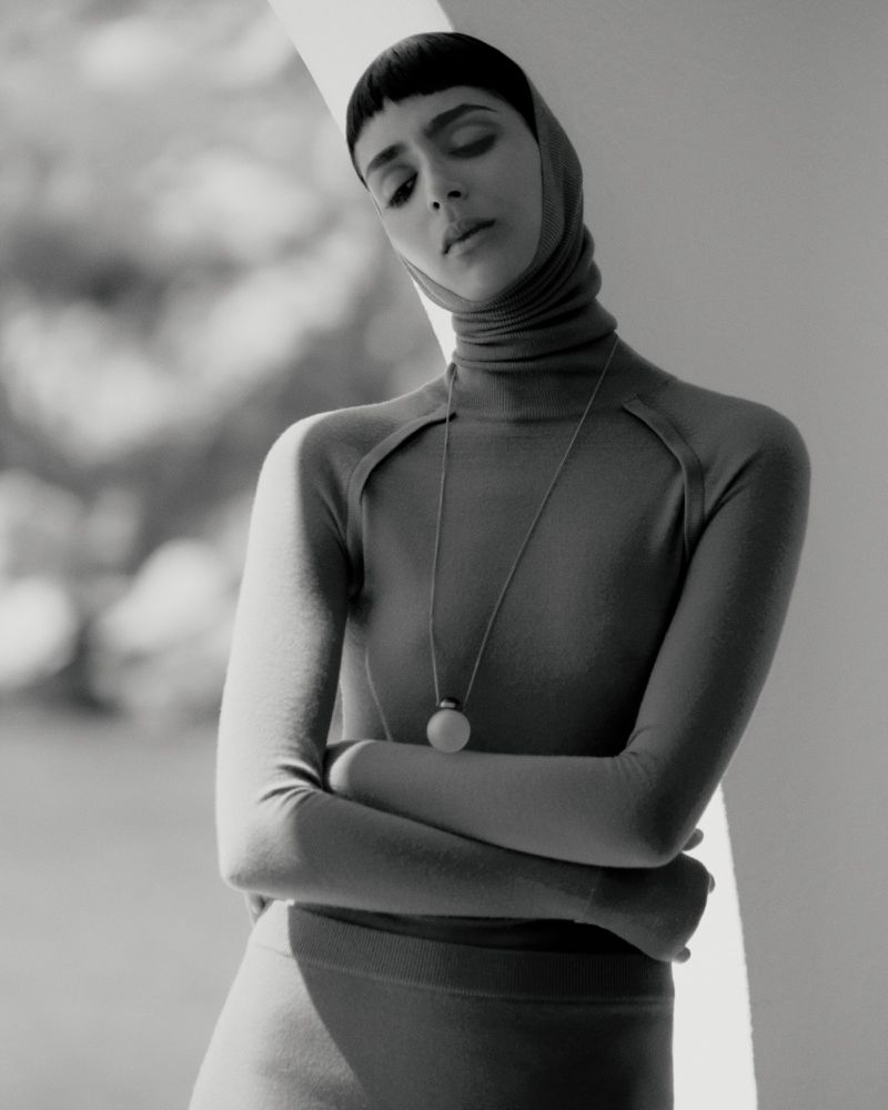 Monochrome portrait of a woman with arms crossed, wearing a turtleneck and pendant necklace in a brightly lit interior space.