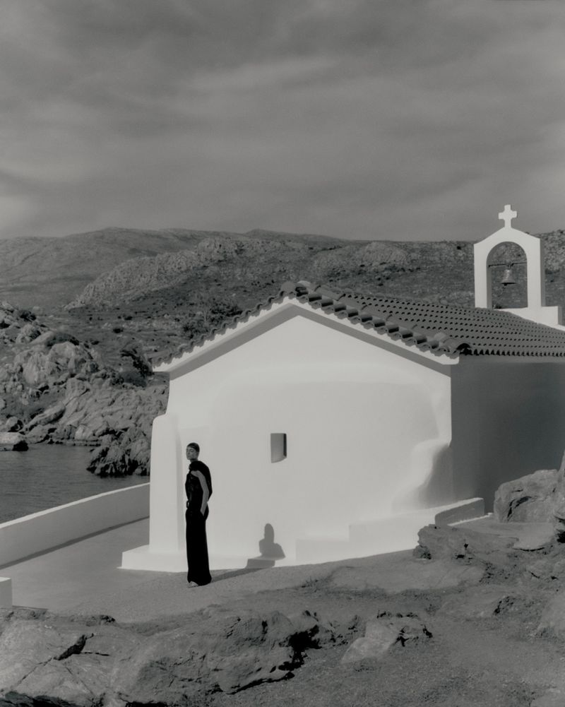 Monochrome shot of an adult in a long dress standing near a building with a cross on the roof and distant rocky hills.