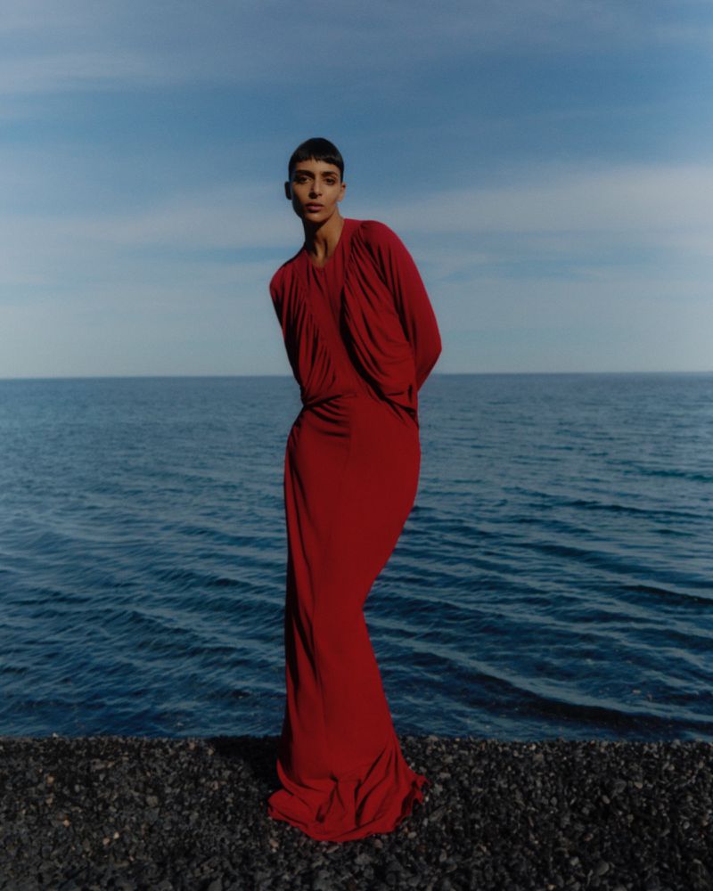 Woman poses in a red gown on a rocky beach by the ocean with a blue sky. The dress has a draped neckline and fitted skirt.