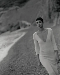 Model Nora Attal walking along a beach in a long deep V-neck knit dress in a black and white fashion editorial.