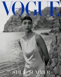 Model Nora Attal in a silver dress posing by a rocky coastal shore for a Vogue Greece magazine editorial cover shot.