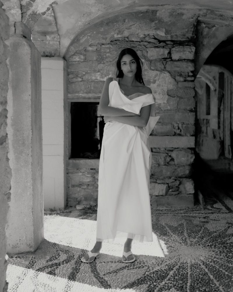 Monochromatic full shot of a woman in a white dress with crossed arms standing in an outdoor stone structure.