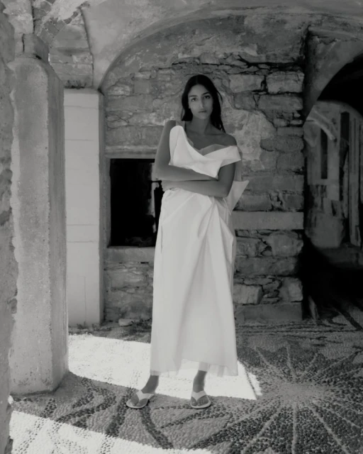 Monochromatic full shot of a woman in a white dress with crossed arms standing in an outdoor stone structure.