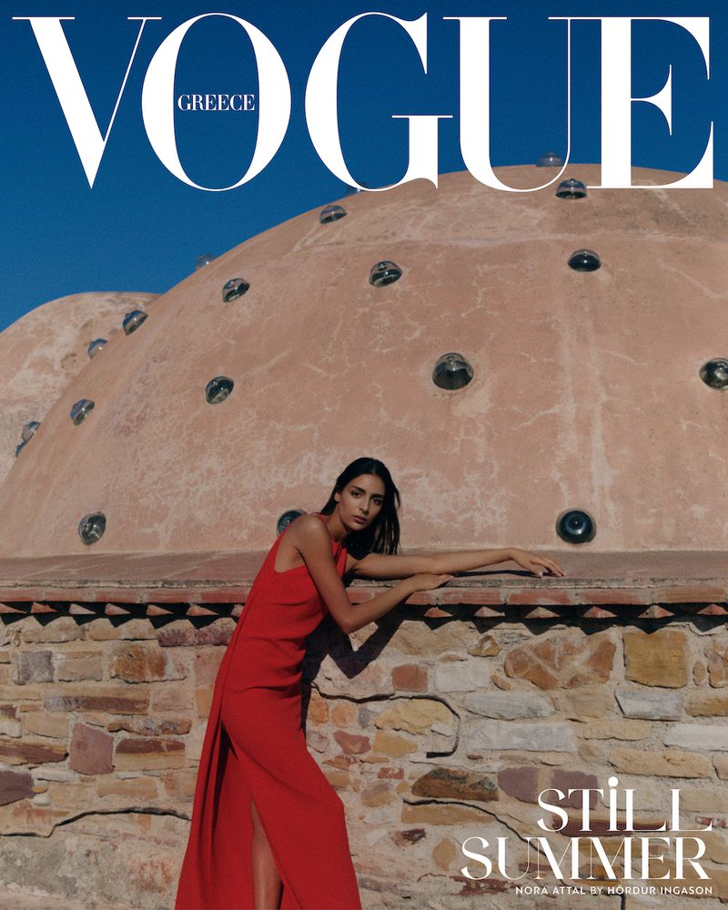 Vogue Greece cover featuring a woman in a red dress leaning against a stone wall with a domed building and blue sky in the background.
