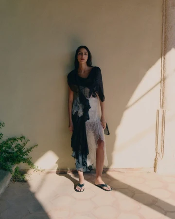 A woman poses in a black and white dress, standing in front of a wall with shadows and green plants, creating a fashion-forward image.