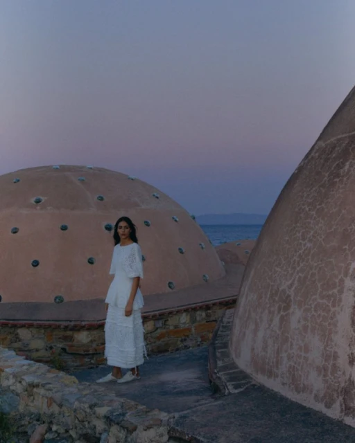 A woman in a white dress stands on a stone platform, domes are behind her, soft dusk lighting gives a sense of serenity by the water.