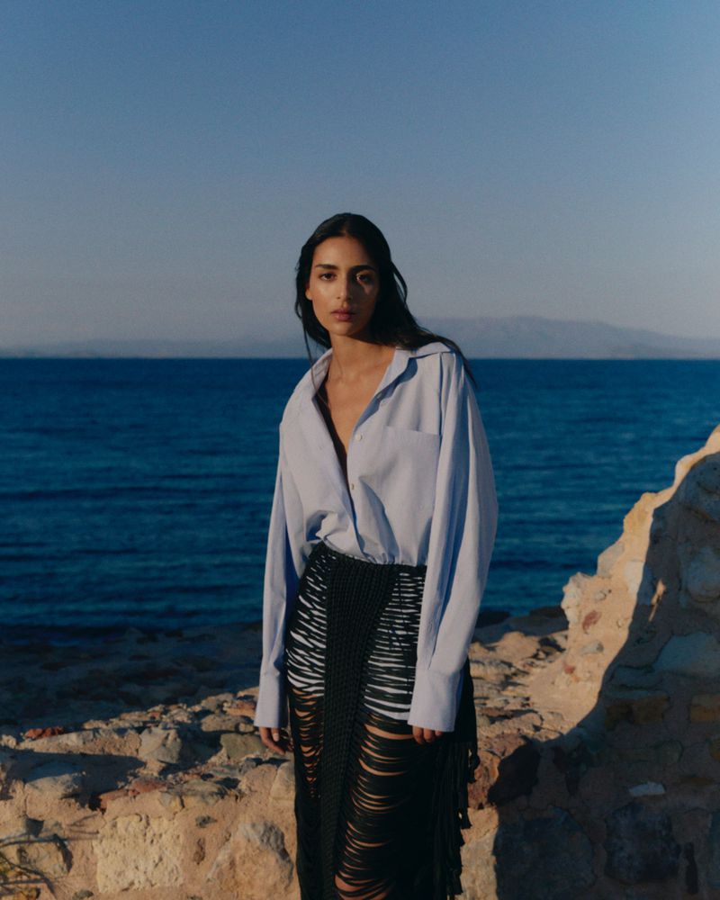 A woman in a light-blue shirt and black fringed skirt poses on a stone ledge overlooking the ocean, mountains on horizon.