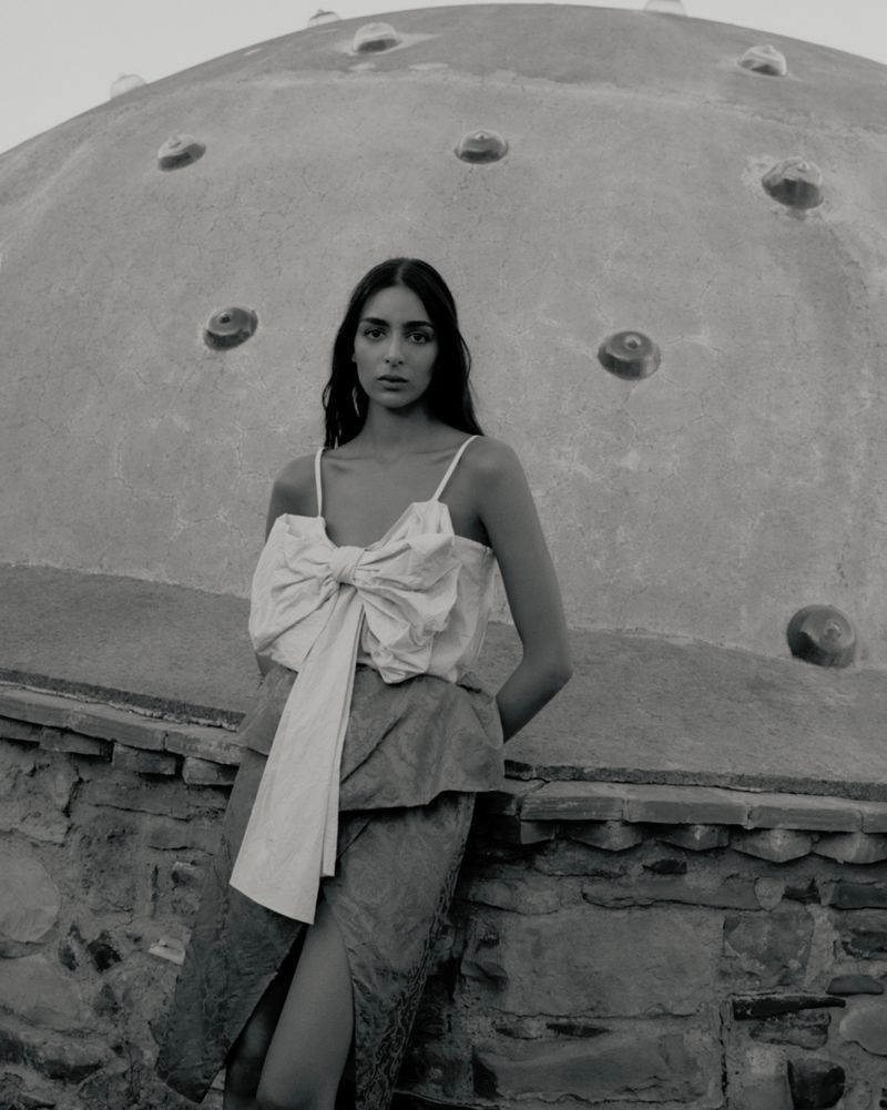 Monochromatic portrait of a woman wearing a ribbon top and skirt, leaning against a stone wall with a domed structure in the background.