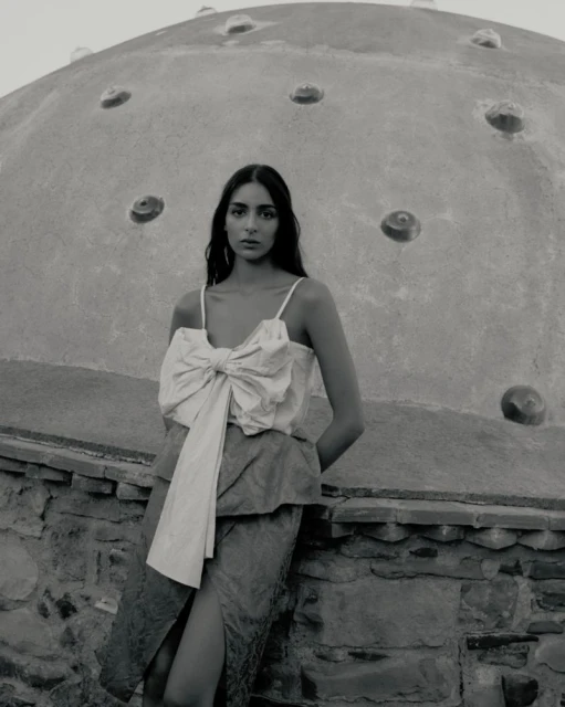 Monochromatic portrait of a woman wearing a ribbon top and skirt, leaning against a stone wall with a domed structure in the background.