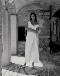 Model Nora Attal standing with arms crossed wearing a white off-shoulder draped gown on a mosaic stone floor.