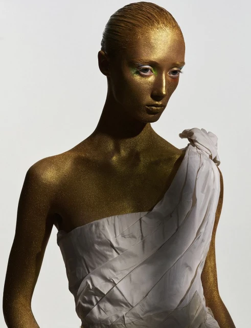 Woman with shimmering gold body paint and white draped fabric, medium shot, against white background in beauty fashion editorial.