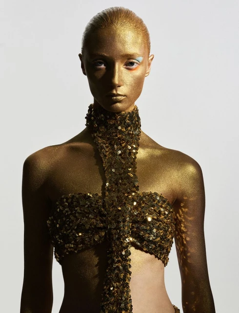 Close-up shot of an adult female covered in gold body paint with orange and blue eye makeup, looking directly at the camera in a photo studio.