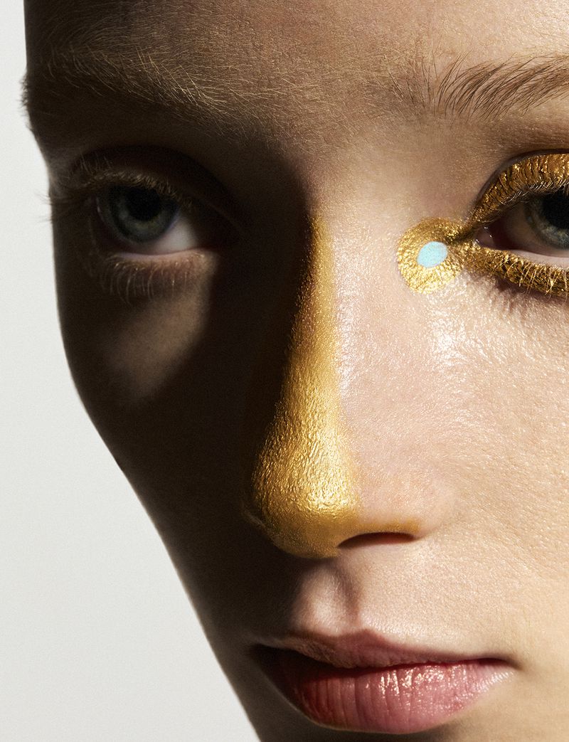 Close-up portrait of a fair-skinned person with gold makeup and dramatic shadows, highlighting eyes and facial structure against a white backdrop.
