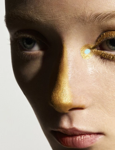 Close-up portrait of a fair-skinned person with gold makeup and dramatic shadows, highlighting eyes and facial structure against a white backdrop.