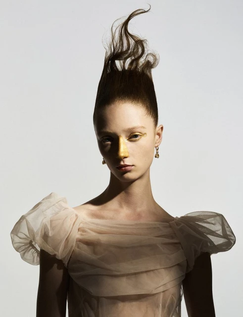 Portrait of a young woman with creative brown hairstyle, golden makeup, beige tulle dress, and drop earrings, posing in a photo studio.