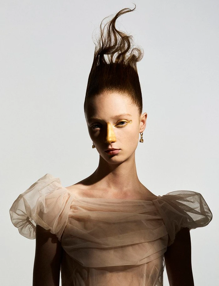 Model Lydia Reid with sculpted swept-up hair and gold nose makeup wearing a sheer ruched tulle top against a plain studio backdrop.